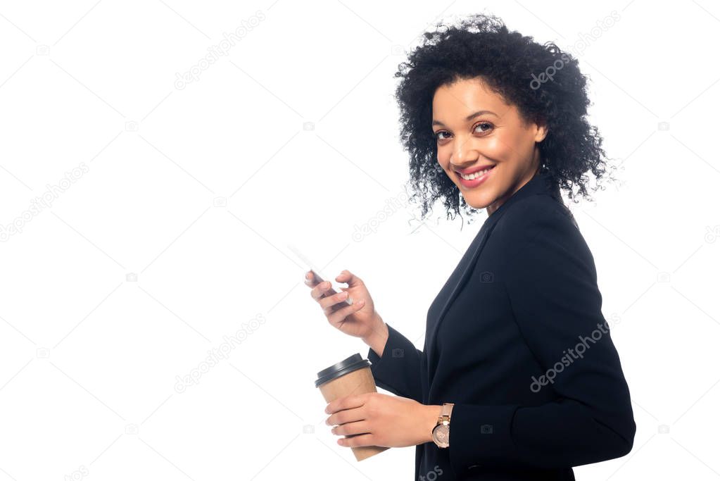 Side view of african american woman smiling with disposable cup of coffee and smartphone isolated on white