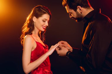 Siyah arka planda, gülümseyen kız arkadaşın parmağına yüzük takan adamın yan görüntüsü.