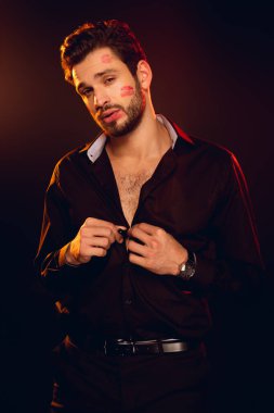 Handsome man with lipstick prints on face looking at camera and unbuttoning shirt isolated on black clipart