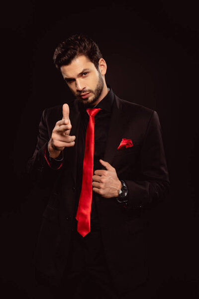 Handsome man in suit pointing with fingers and looking at camera isolated on black