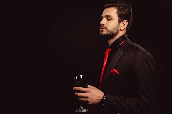 Side view of elegant man in suit holding glass of red wine isolated on black