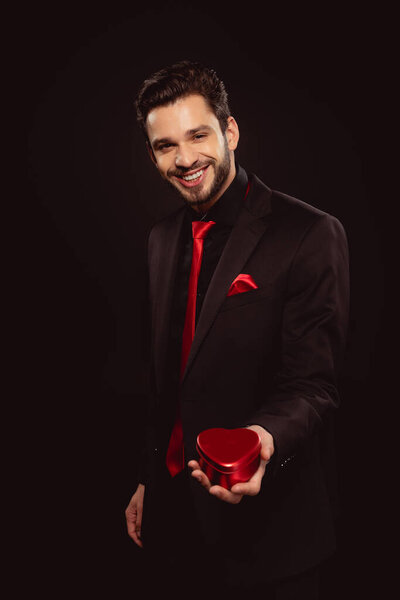 Smiling man in formal wear holding gift box in heart shape and looking at camera isolated on black