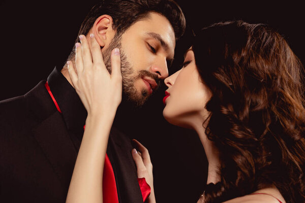 Side view of beautiful woman kissing and touching face of elegant boyfriend isolated on black