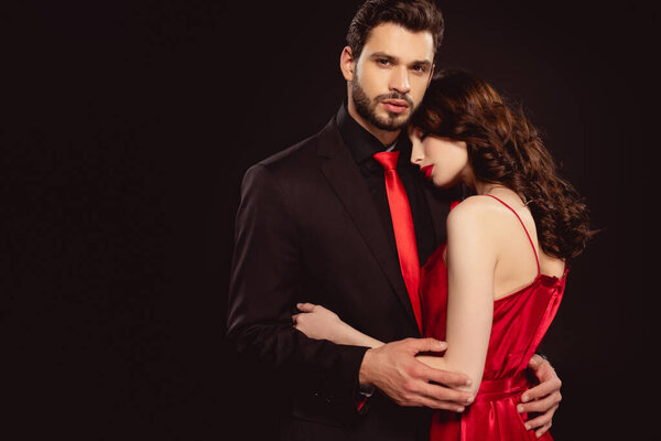 Handsome man embracing beautiful woman in red dress and looking at camera isolated on black