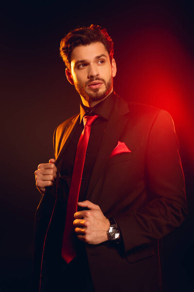 Handsome man looking away while adjusting jacket on black background with lighting