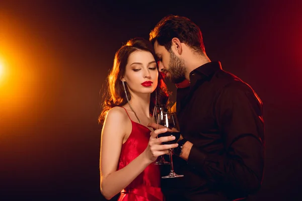 一个英俊的男人在优雅的女朋友身边举着一杯红酒 背景是黑色的 点着灯 — 图库照片