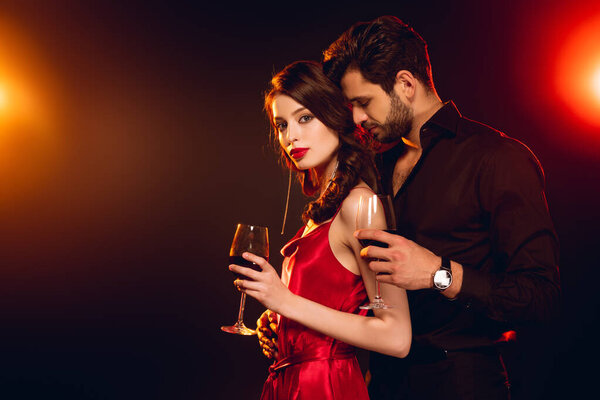 Side view of woman holding glass of wine near elegant boyfriend on black background with lighting