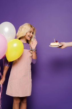 Mor arka planda balonları olan gülümseyen çocuğa doğum günü pastası veren bir kadın manzarası. 