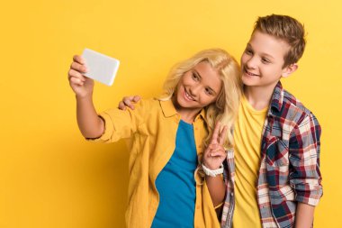 Gülümseyen çocuk barış hareketi yapıyor ve sarı arka planda arkadaşıyla selfie çekiyor. 