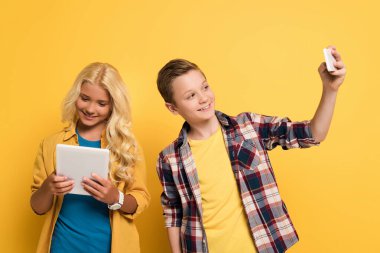 Gülümseyen çocuk selfie çekiyor ve arkadaşı sarı arka planda dijital tablet kullanıyor. 