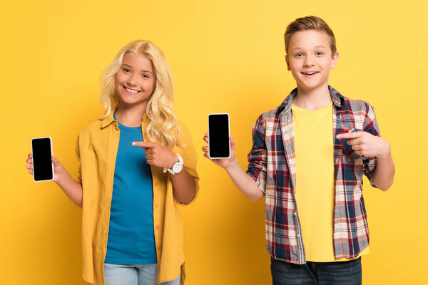 улыбающиеся дети, указывающие пальцами на смартфоны на желтом фоне
 