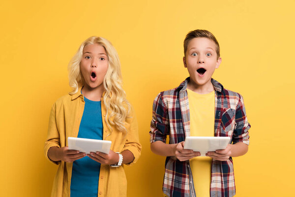 shocked kids holding digital tablets and looking at camera on yellow background 