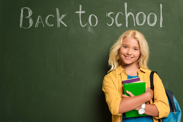 smiling schoolgirl with books standing near chalkboard with back to school lettering