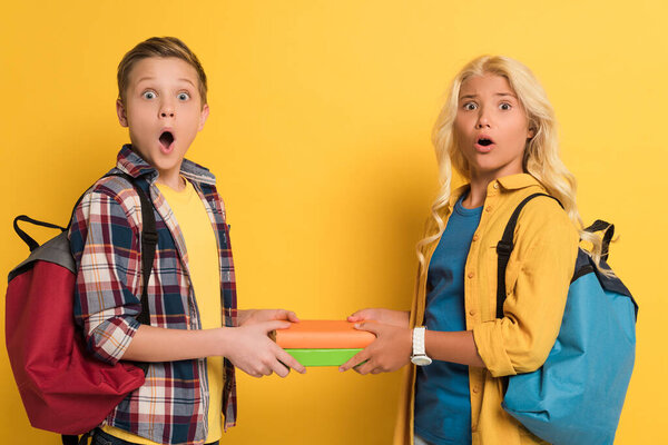 shocked schoolkids holding books and looking at camera on yellow background 