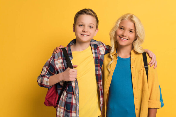 smiling schoolkids with backpacks hugging and looking at camera on yellow background 