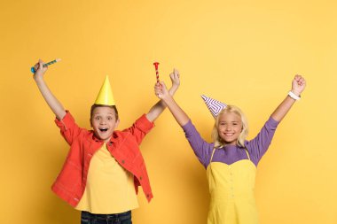 Sarı arka planda parti boynuzları tutan elleri açık gülümseyen çocuklar. 