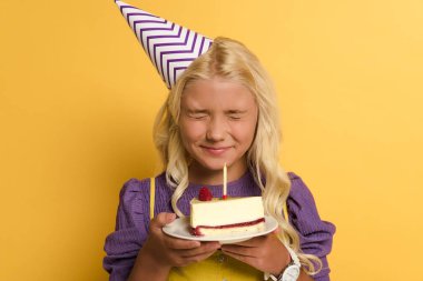 smiling kid holding plate with birthday cake and making wish on yellow background  clipart