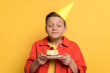 smiling kid with closed eyes making wish and holding plate with birthday cake on yellow background  clipart