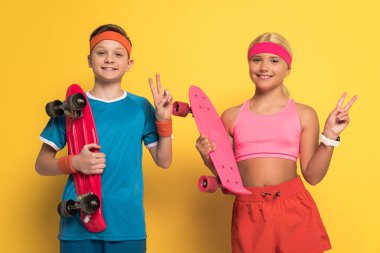 Spor kıyafetleri içinde gülümseyen çocuklar ellerinde bozuk paralarla sarı arka planda barış jestleri gösteriyorlar. 