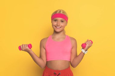 Sarı arka planda dambıllar ile gülümseyen genç bayan sporcu eğitimi 