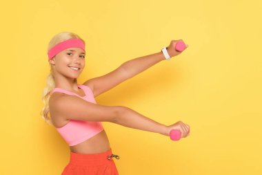 Sarı arka planda dambıllar ile gülümseyen genç bayan sporcu eğitiminin yan görüntüsü 