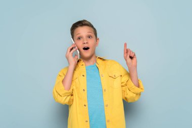 Şaşırmış çocuk akıllı telefondan konuşuyor ve mavi arka planda fikir jesti gösteriyor. 