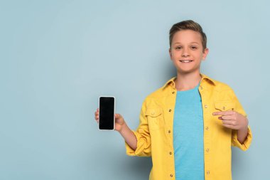 Gülümseyen çocuk mavi arka planda akıllı telefonu işaret ediyor. 