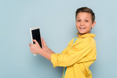 Elinde dijital tablet olan gülümseyen çocuk mavi arka plandaki kameraya bakıyor. 