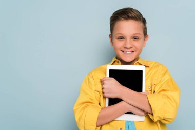 Elinde dijital tablet olan gülümseyen çocuk mavi arka plandaki kameraya bakıyor. 