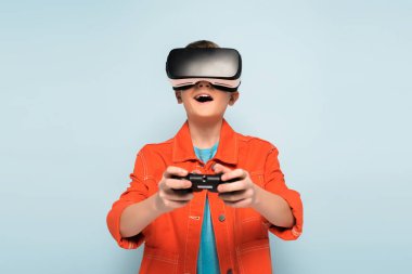 KYIV, UKRAINE - 21 HAZİRAN 2020: Sanal gerçeklik kulaklığı olan şok olmuş çocuk mavi arka planda joystick ile oynuyor 