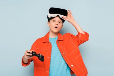 KYIV, UKRAINE - 21 Ocak 2020: Sanal gerçeklik kulaklığı olan şok olmuş çocuk mavi arka planda joystick tutuyor 
