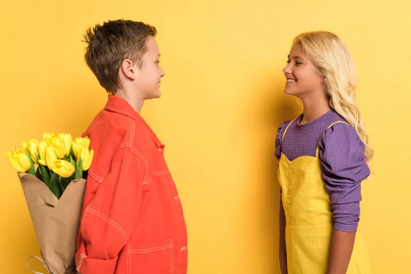 Sarı Arka Planda Sevimli Arkadaşından Buket Saklayan Gülümseyen Çocuğun Yan — Stok fotoğraf