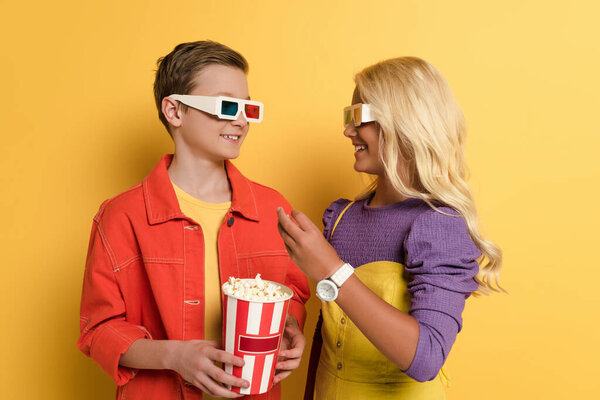 smiling kids with 3d glasses holding popcorn and looking at each other on yellow background 