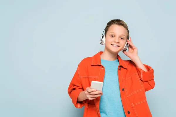 smiling kid with headphones listening to music and holding smartphone on blue background