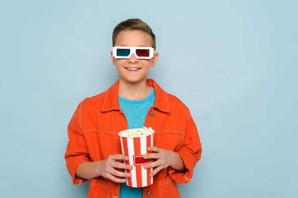 улыбающийся ребенок в 3D очках, держащий ведро с попкорном на синем фоне
 