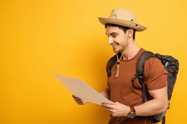 handsome smiling tourist in hat with backpack looking at map on yellow clipart