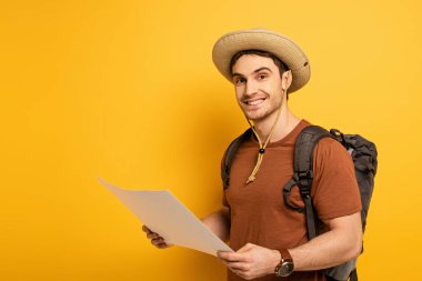 handsome smiling tourist in hat with backpack holding map on yellow clipart