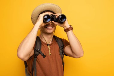 smiling traveler in hat with backpack looking through binoculars  on yellow clipart
