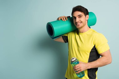 Sarı tişörtlü gülümseyen kaslı sporcu spor paspası ve mavi üzerine su dökülmüş spor şişesi tutuyor.