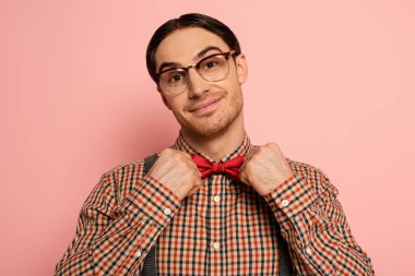 smiling male nerd in eyeglasses adjusting bow tie on pink clipart