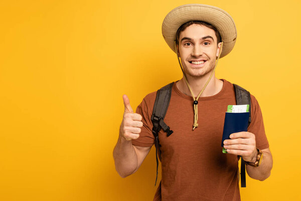 cheerful tourist in hat with backpack holding passport and ticket while showing thumb up on yellow