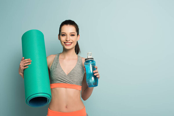 smiling sportswoman holding fitness mat and sports bottle with water on blue