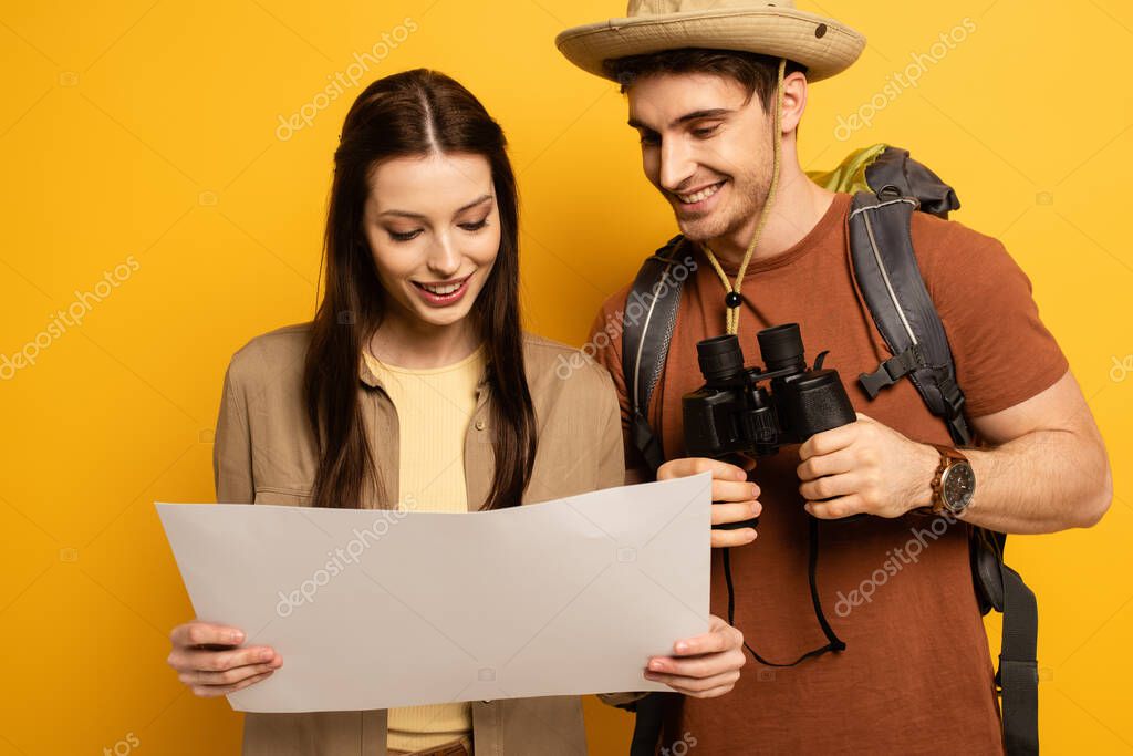couple of smiling travelers with backpack and binoculars looking at map on yellow