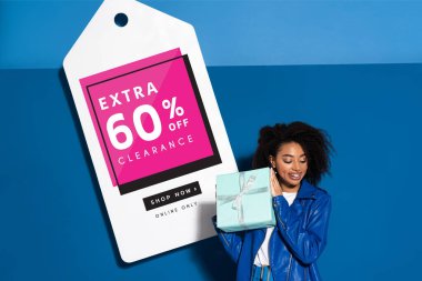 smiling african american woman with gift near big price tag with extra 60 percent off clearance illustration on blue background