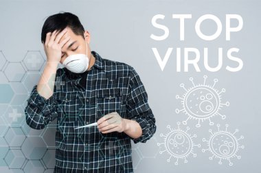 Solunum maskeli genç Asyalı adam termometreye bakıyor ve gri arka planda alnına dokunuyor. Virüs çizimini durdur.