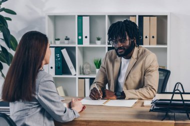 African american recruiter with pen and notebook conducting job interview with employee in office clipart