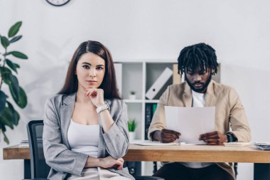 African american recruiter reading documents with employee looking at camera at table in office clipart