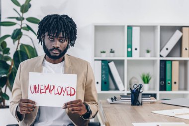 Afro-amerikan çalışan, ofisinde işsiz harfleri olan bir pankart gösteriyor.