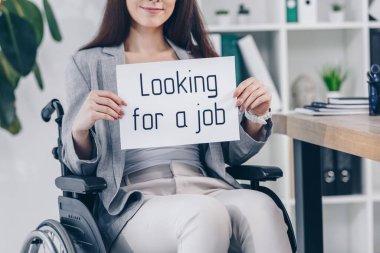 Cropped view of disabled employee smiling and holding placard with looking for a job lettering on wheelchair in office clipart