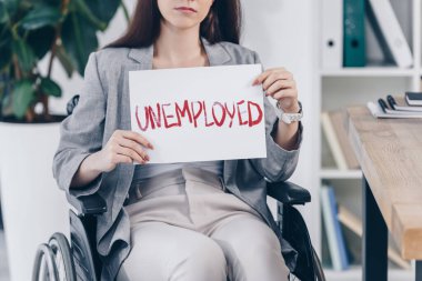 Tekerlekli sandalyede işsiz harfleriyle pankart tutan engelli bir çalışanın görüntüsü.